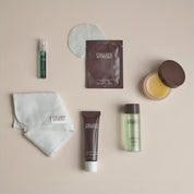 A flat-lay arrangement of six Collagen Boosted skincare products on a warm beige background, including a white cotton pad, a small green spray bottle, a brown sachet, a folded white cloth, a dark brown tube, a clear bottle with pale green liquid, and a jar of yellow balm.