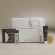 Colleen Rothschild The Skincare Kit arranged on a neutral beige surface with a soft beige backdrop. The set includes a large white gift box with a folded white towel draped over the top, a dark brown sachet, a pale green bottle, a dark brown tube, a small clear spray vial, and a round clear container holding a yellow balm.