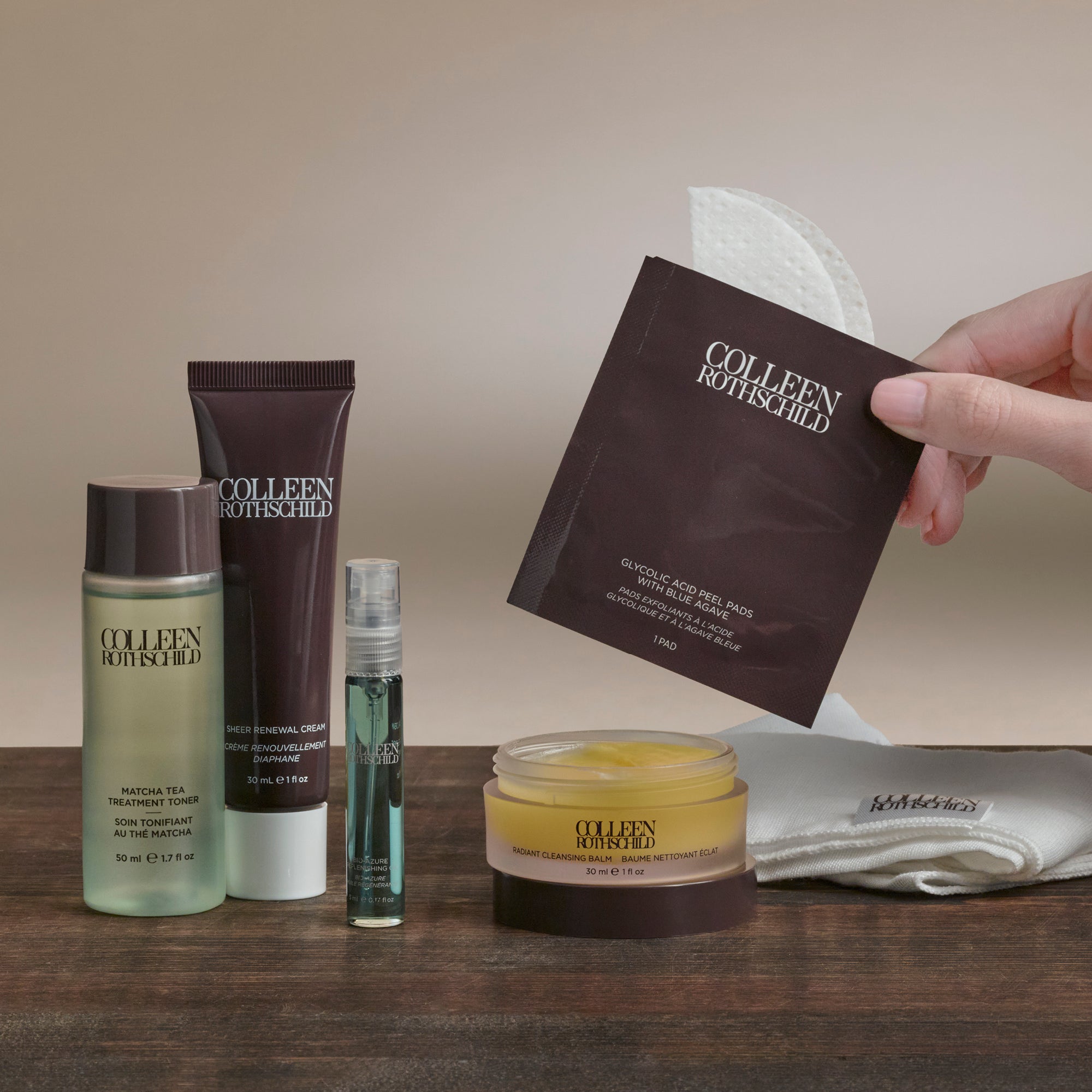 A hand holds a brown COLLEEN BOUTIQUE facial mask sachet over a styled collection of COLLEEN BOUTIQUE skincare products on a wooden table, including a toner, cream, serum, and a jar of facial mask cream, with a white towel in the background.