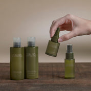 Four Colleen Matthews haircare bottles in olive green and translucent shades, arranged on a wooden surface with a hand holding one bottle above them against a warm beige background.