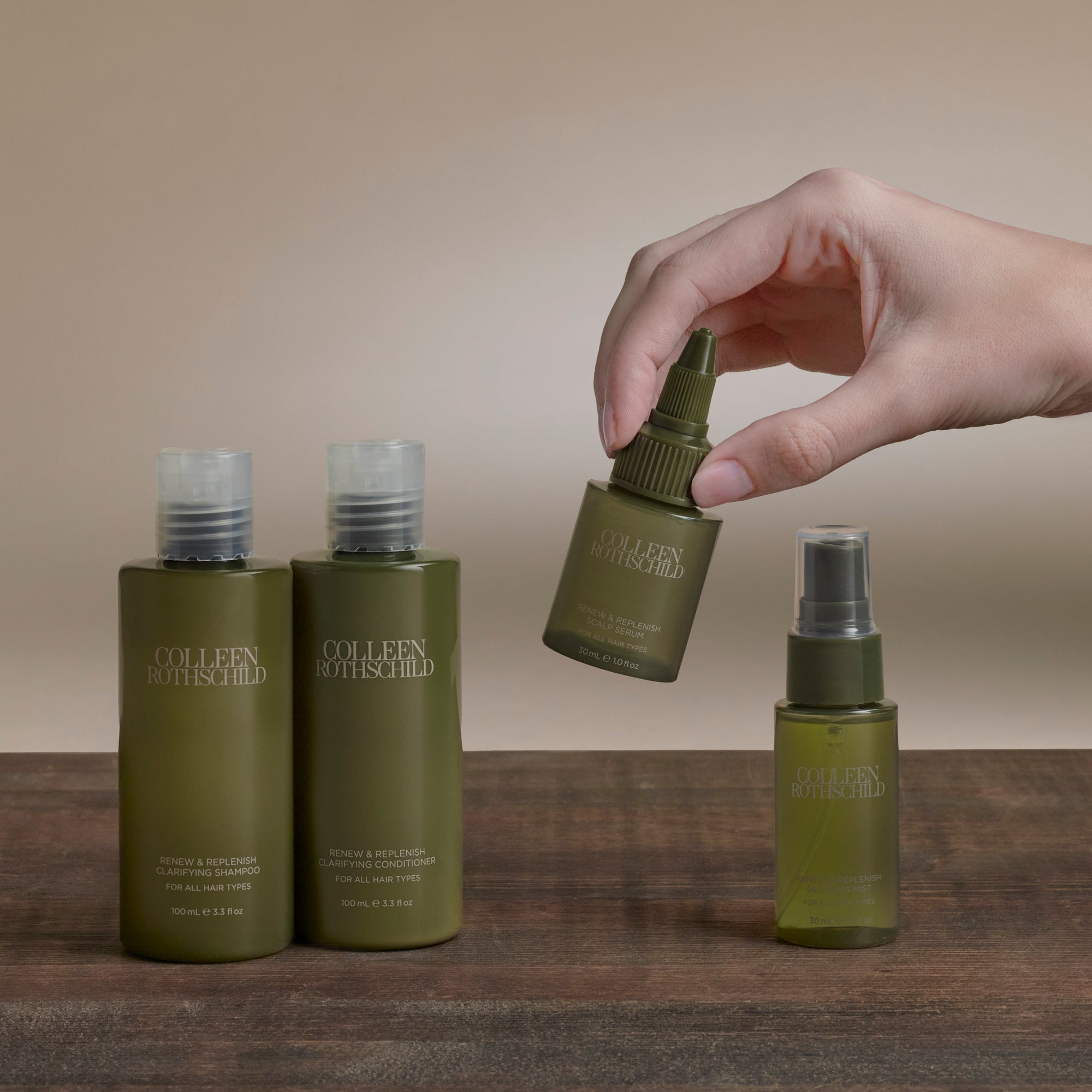 Four Colleen Matthews haircare bottles in olive green and translucent shades, arranged on a wooden surface with a hand holding one bottle above them against a warm beige background.