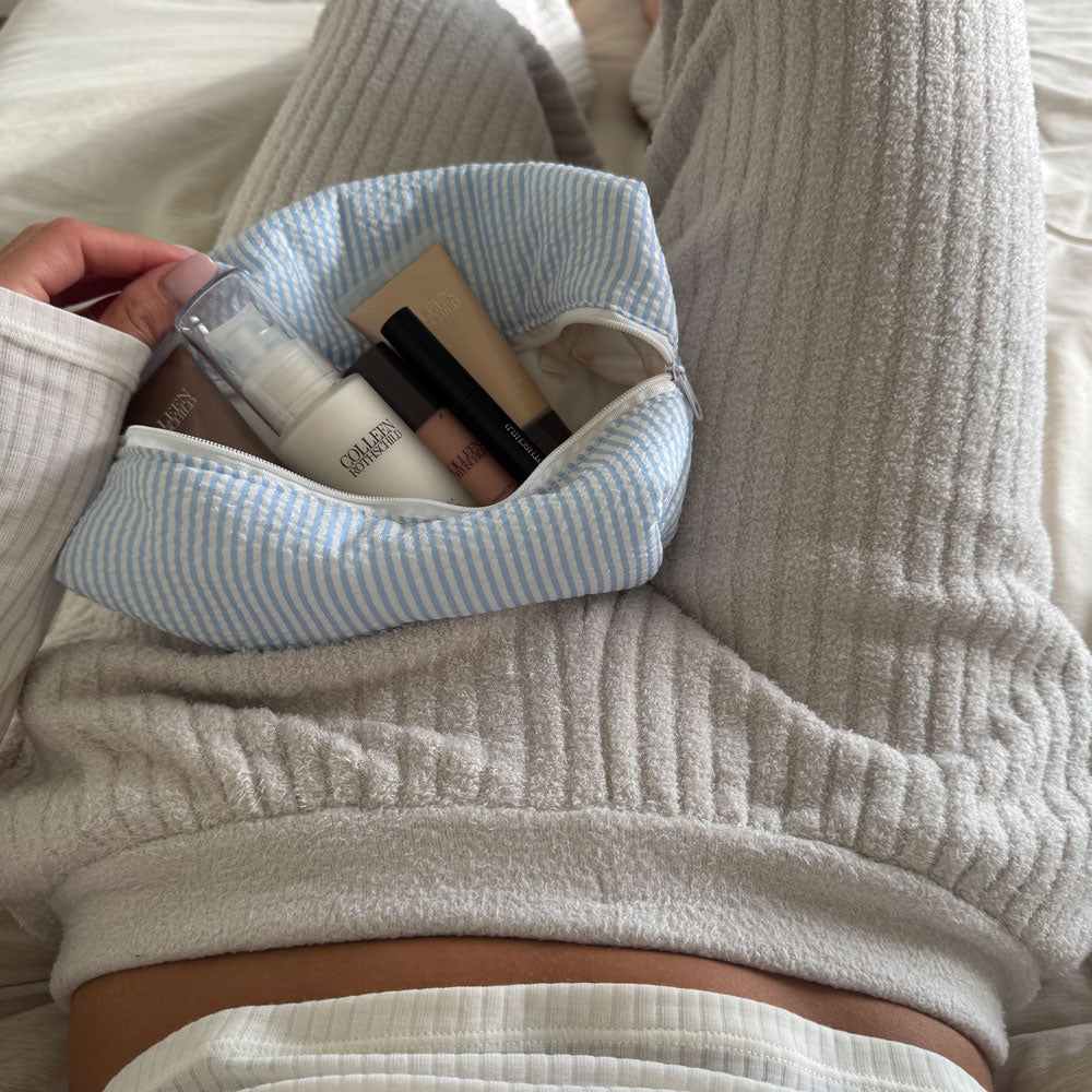 Light blue striped makeup bag with Colleen Rothschild skincare and makeup items held in a woman's lap.